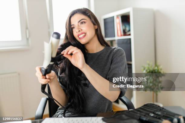 frau schaut auf computer und mit ihrem lockenbügeleisen, während sie am schminktisch sitzt. frau macht ihre haare, indem sie ein tutorial im internet zum zeitpunkt der corona-virus-pandemie - lockenstab stock-fotos und bilder
