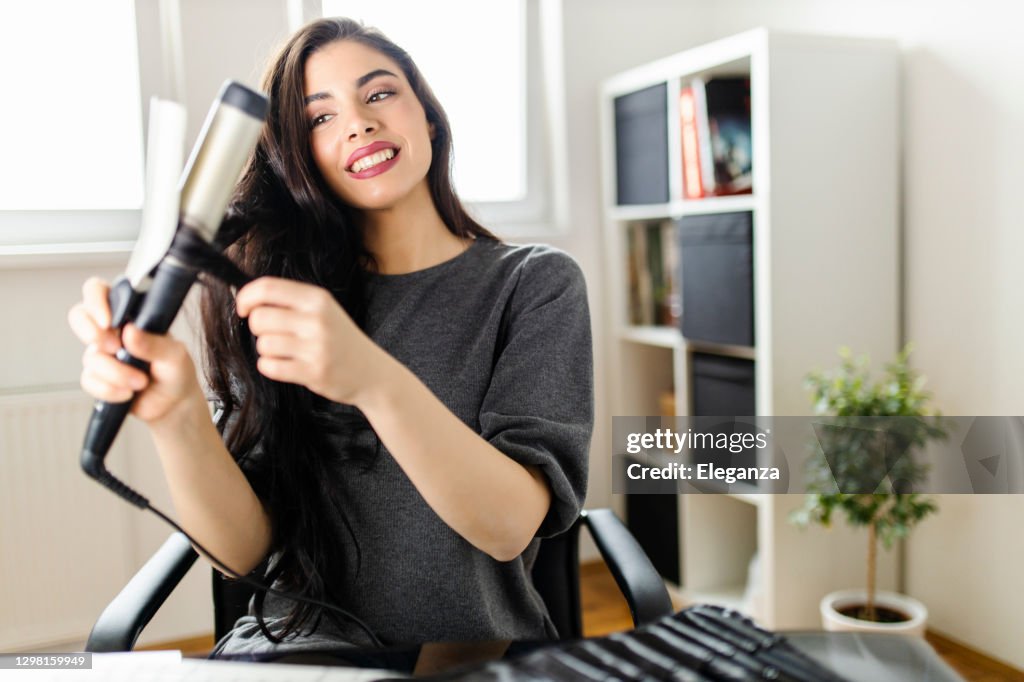 Vrouw die computer bekijkt en haar krultang gebruikt terwijl het zitten bij de kaptafel. Vrouw doet haar haar door te kijken naar een tutorial op het internet op het moment van de corona virus pandemie