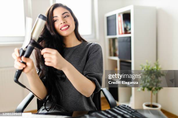 woman looking at computer and using her curling iron while sitting at the dressing table. woman does her hair by watching a tutorial on the internet at the time of the corona virus pandemic - hair curlers stock pictures, royalty-free photos & images