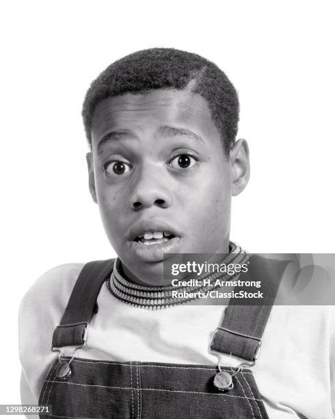 1970s Surprised African-American Pre-Teen Boy Wearing T-Shirt Bib Overhauls Wide-Eyed Looking At Camera.