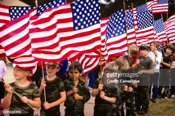 1980s Group Of Patriotic Boys Wearing Camouflage Pattern T-Shirts Carrying American Flags Attending National Celebration Parade.