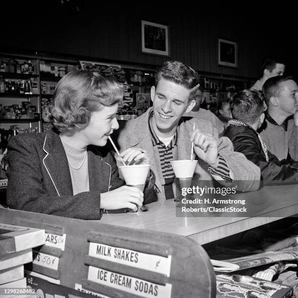 1950s Teenage Couple Boy And Girl Sitting Together Drinking Soft Drinks At Soda Fountain Counter Laughing Smiling Flirting.