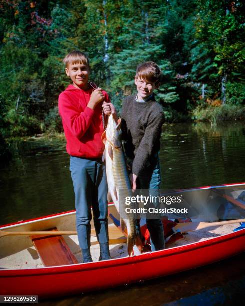 1960s Two Smiling Boys Brothers Standing In Red Canoe Looking At Camera Holding Up Big Fish Ballard Lake Northern Wisconsin USA.