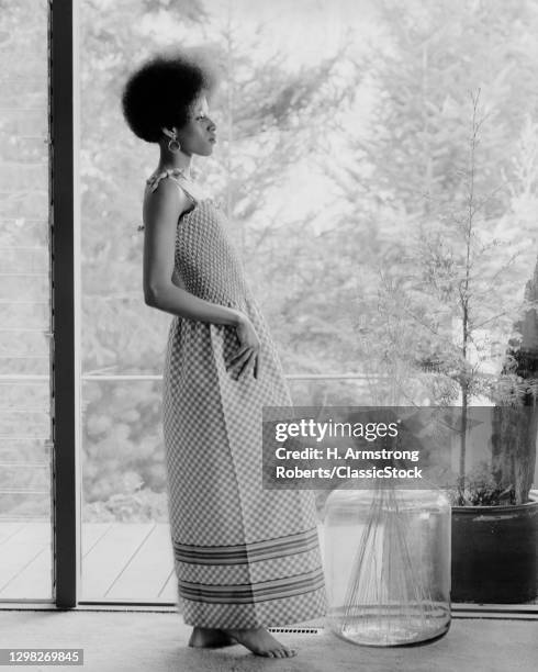 1970s African American Woman Afro Hairstyle Barefoot In Fashionable Maxi Dress With Smocked Bodice Before Standing A Window.