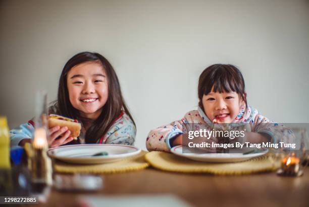 lovely little sibling smiling joyfully at the camera while enjoying breakfast at home in the morning - asian eating hotdog stock pictures, royalty-free photos & images