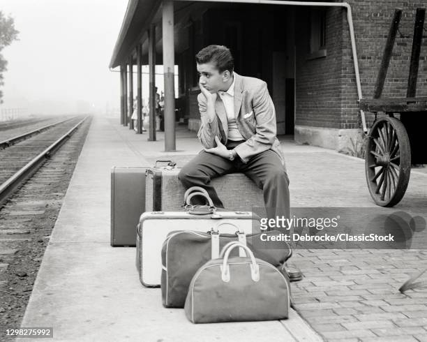 1950s Teen Boy Sitting On Luggage Train Station Platform Waiting For Train To Arrive Sad Expression Leaving Going Back To School.