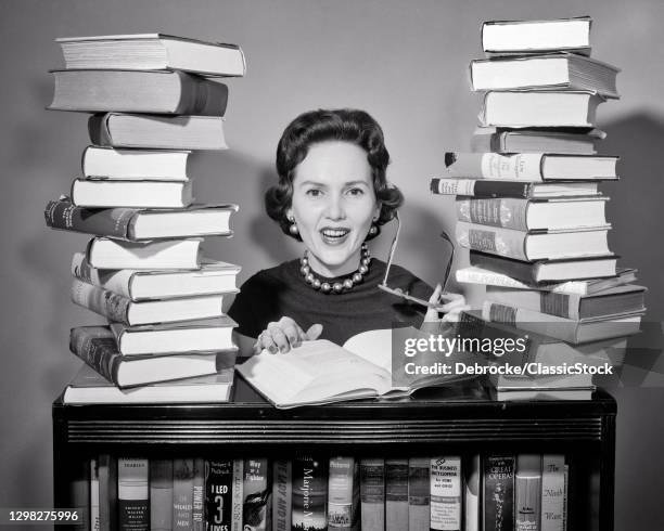 1950s Smiling Woman Teacher Librarian Behind A Book Case Stacks Of Books Beside Her Holding Eyeglasses Looking At Camera .