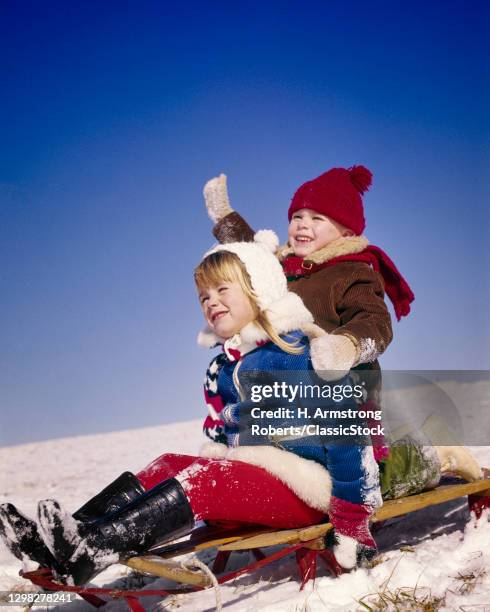 1970s Boy And Girl Blonde Smiling Laughing Wearing Winter Clothes Gloves Boots Hats Sled Riding Down Snowy Hill Boy Waving Arm.