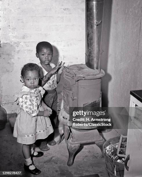 1950s 1960s Two Young African American Children Boy Girl Looking At Camera Keeping Warm By Cast Iron Stove Poor Welfare Housing.