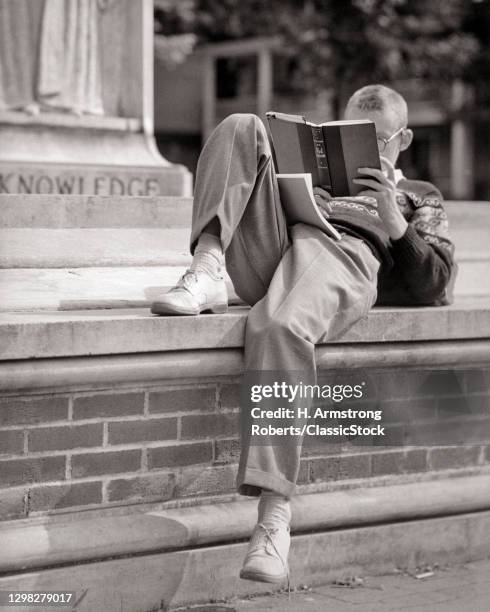 1950s Anonymous Blonde Boy Higher Education Student Sitting Reclining On Brick Wall Campus Steps Engrossed In Reading A Book.