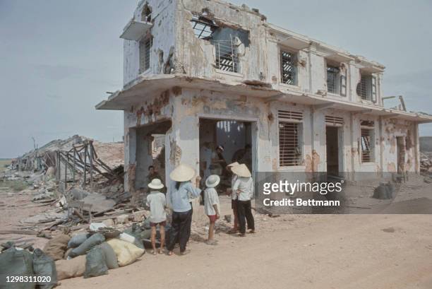 Villagers, some wearing a traditional non la hats, look on as South Vietnamese troops search a bombed-out building along Highway 555 some four...