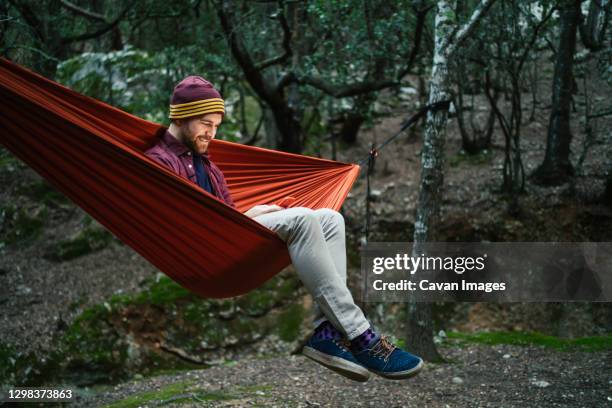 young male uses his smartphone resting on a hammock in the woods - male cellphone stock-fotos und bilder