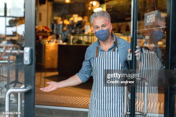 business owner wearing a facemask while welcoming customers to his cafe - open sign on door stock pictures, royalty-free photos & images