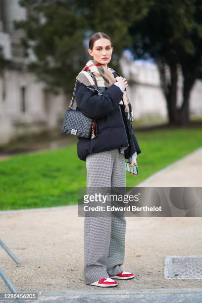 Model wears a checked plaid wool scarf, a black winter puffer jacket, a black leather quilted Chanel bag, gray flared pants, red and white sneakers...