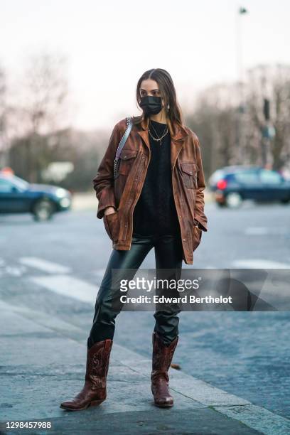 Model wears a black protective face mask, a necklace, a brown leather jacket, a black pullover, black leather pants, brown leather cowboy boots,...