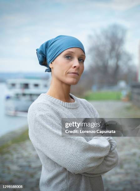 young woman with cancer, wearing a blue bandana, looks at the camera, with a determined and confident expression, and with her arms crossed. - cancer stock-fotos und bilder