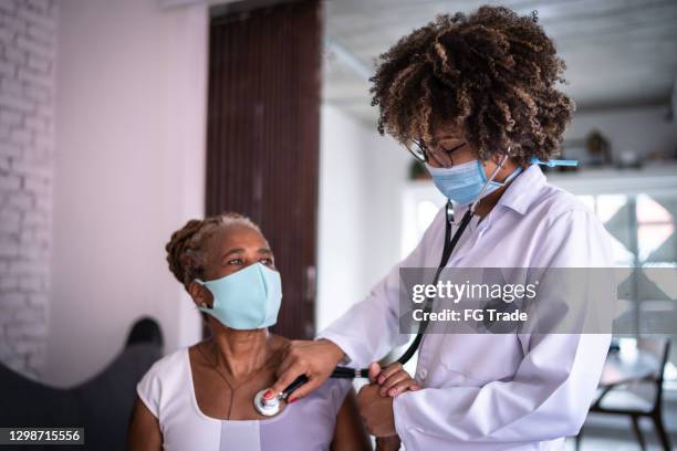 medico che utilizza il battito cardiaco del paziente anziano in ascolto durante una visita a casa - usando una maschera facciale - servizio a domicilio foto e immagini stock