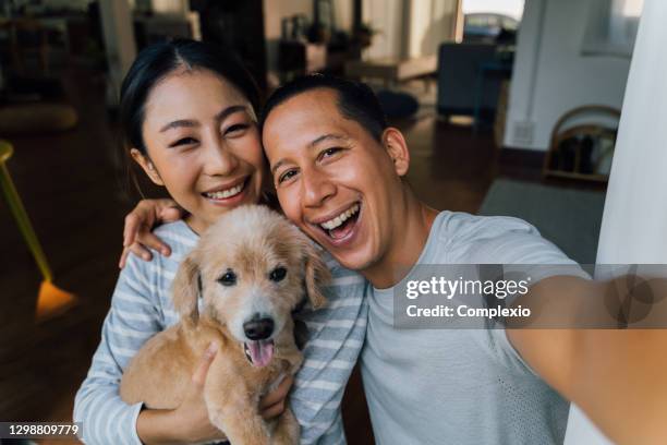 young adult asian couple holding a puppy taking a selfie from a phone with home interior in background. 30s mature man and woman with dog pet taking a family photo shots. - happy group portrait. - family zoom stock pictures, royalty-free photos & images