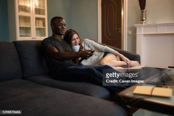diverse couple cuddling on sofa and watching tv together - male looking content stock pictures, royalty-free photos & images