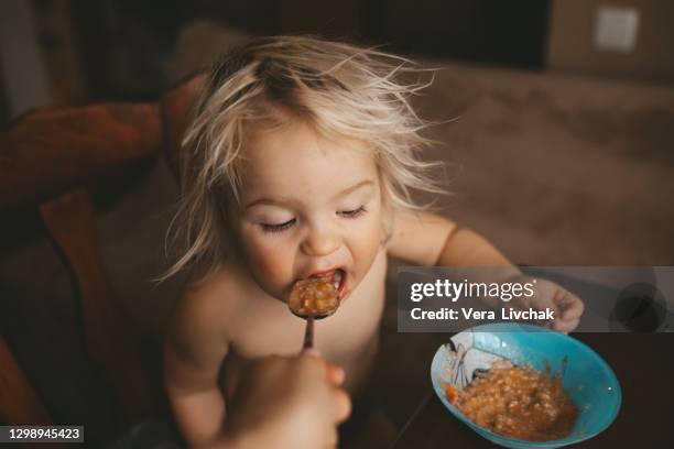 portrait of 2 years old baby boy sitting in chair and eatting porridge from spoon - funny baby faces stock pictures, royalty-free photos & images