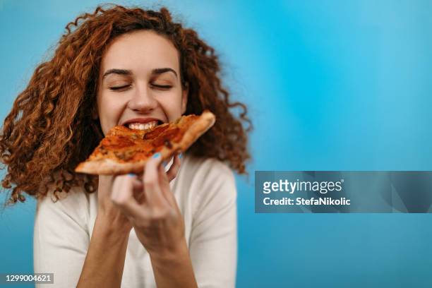 woman standing in front of blue background and eating pizza - pizza imagens e fotografias de stock