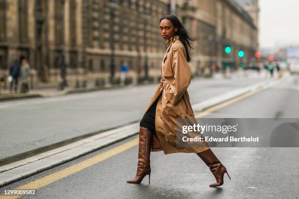 Emilie Joseph wears a golden large chain necklace from JW Anderson, a beige pale brown long trench coat from Gestuz, knee high brown leather boots...