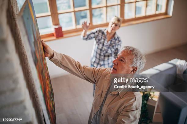 senior couple in their new home hanging a painting together - hanging stock pictures, royalty-free photos & images