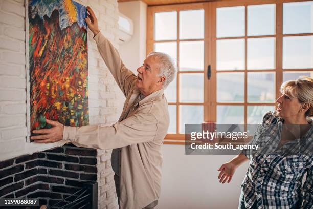 senior couple in their new home hanging a painting together - house for an art lover stock pictures, royalty-free photos & images
