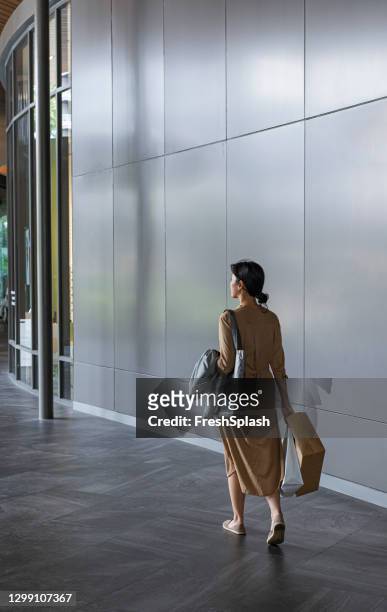rückblick auf eine junge asiatin, die in einem einkaufszentrum mit einkaufstaschen in den händen spazieren geht - graues kleid stock-fotos und bilder
