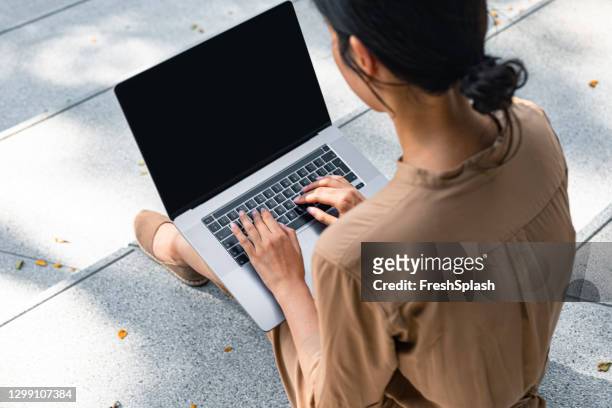 digital nomads: over the shoulder view of a woman sitting on stairs and working online on a laptop pc with a blank screen (copy space) - over the shoulder view stock pictures, royalty-free photos & images