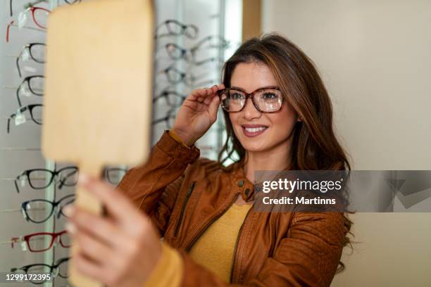 woman choosing glasses in optical store using mirror - choosing eyeglasses stock pictures, royalty-free photos & images