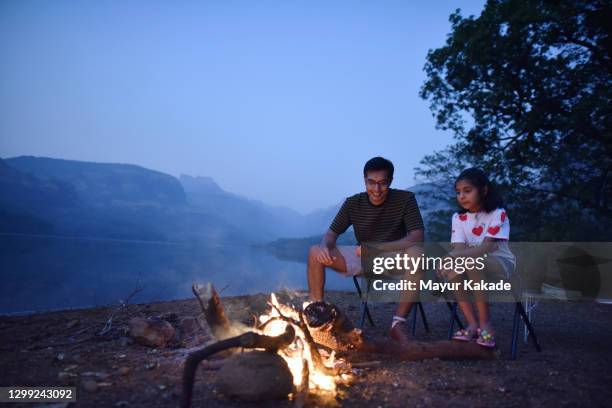 father and daughter sitting near a campfire on chairs by the beautiful lake - indian family vacation stock pictures, royalty-free photos & images