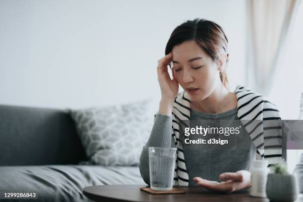 young asian woman feeling sick and holding her head in pain, taking medicines in hand, with a glass of water and pill bottle by the side on coffee table. medicine, healthcare and people concept - headache stock-fotos und bilder
