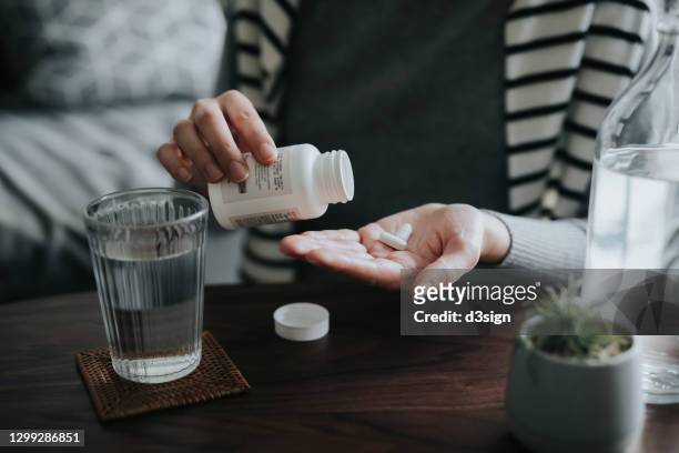close up of young asian woman holding a pill bottle, pouring pills into palm of hand, with a glass of water on the side. medicine, healthcare and people concept - antibiotikum stock-fotos und bilder