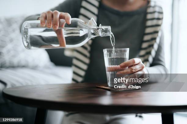 close up of young asian woman pouring water from bottle into the glass on a coffee table at home. healthy lifestyle and stay hydrated - wasser stock-fotos und bilder