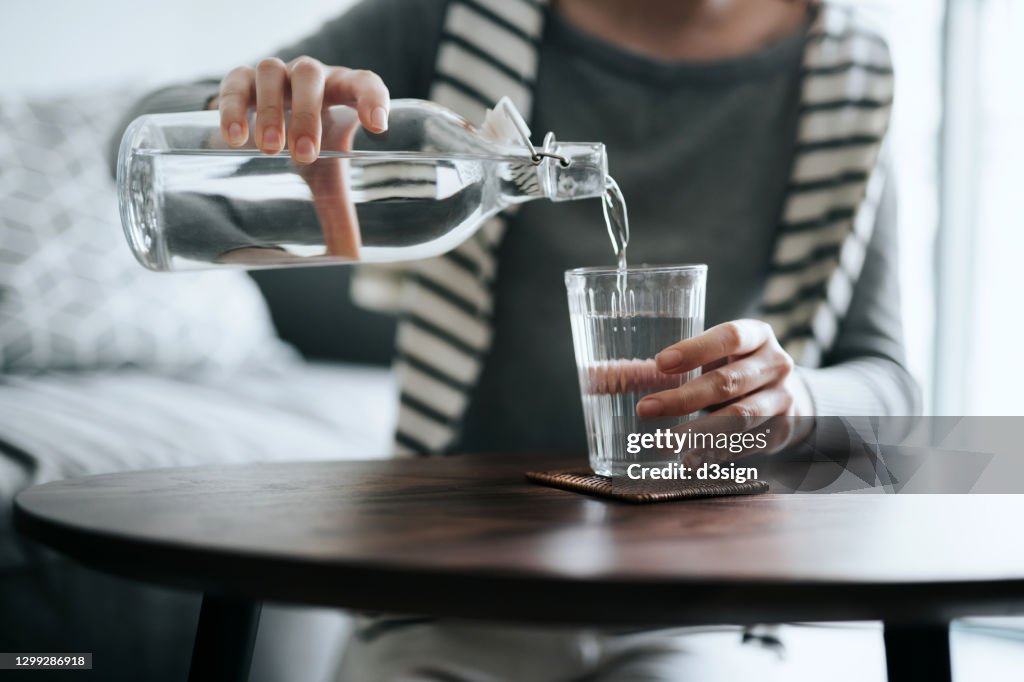Close up of young Asian woman pouring water from bottle into the glass on a coffee table at home. Healthy lifestyle and stay hydrated