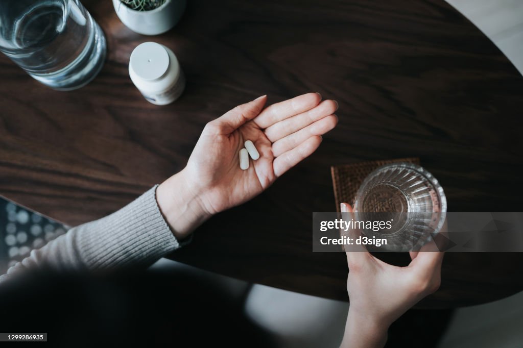 Overhead view of young woman holding a glass of water, taking medicines at home. Medicine, healthcare and people concept