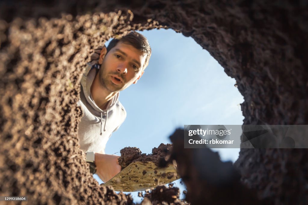 A young guy digging a deep hole with a shovel is surprised by an amazing find. View from the underground at the grave digger