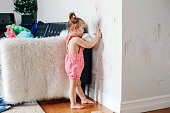 Funny cute baby girl drawing with marker on wall at home. Toddler girl child with milk bottle playing at home. Authentic candid childhood lifestyle moment. Young artist painting on wall at living room