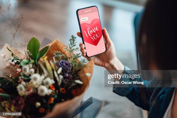 over-the-shoulder view of a unrecognisable woman receiving flower bouquet and reading "i love you" message on mobile phone - quedarse en casa frase fotografías e imágenes de stock