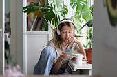 Woman listening to music wear wireless white headphones using mobile smart phone, drinks tea at home