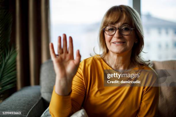 smiling senior woman waving while looking at camera at home - stay at home saying stock pictures, royalty-free photos & images