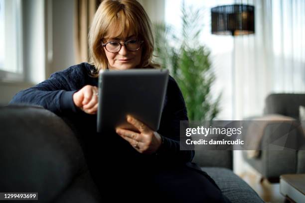 hogere vrouwenzitting op bank en gebruikend digitale tablet thuis - isolated women serieus sad stockfoto's en -beelden