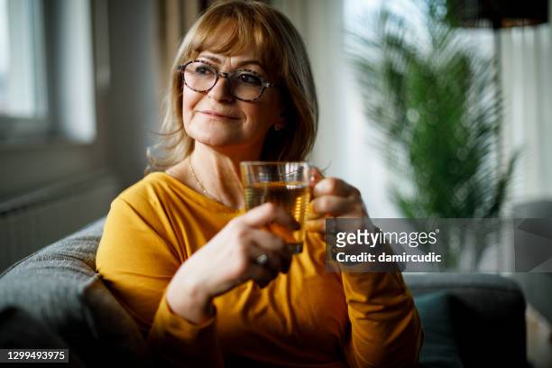 portrait of beautiful smiling senior woman sitting on sofa and drinking tea at home - drinking cold drink stock pictures, royalty-free photos & images