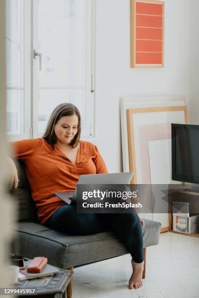 mujer de tamaño más sonriente sentada cómodamente en un sillón y trabajando en un ordenador portátil - voluptuoso fotografías e imágenes de stock
