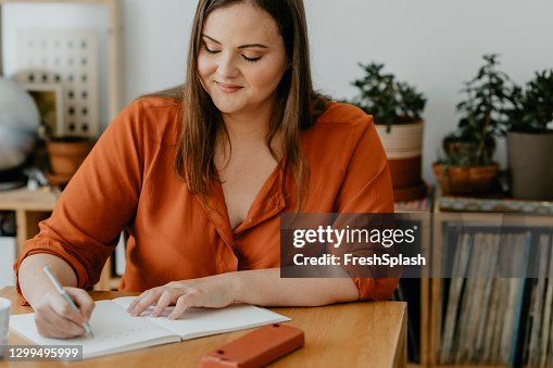 Belle femme en surpoids s’asseyant dans son salon et écrivant un journal Belle femme en surpoids s’asseyant dans son salon et écrivant un journal