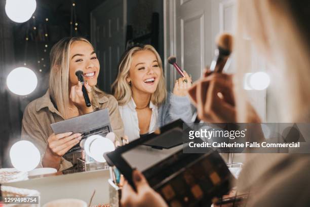 two young woman sit in front of an illuminated mirror and apply make-up - trucco per il viso foto e immagini stock