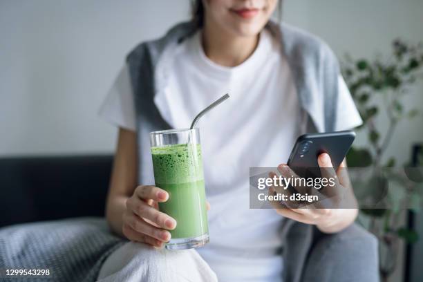 cropped shot of young asian woman using smartphone while sitting on the sofa at home, drinking a glass of fresh green detox juice with reusable metal straw. healthy eating, green colour and detox diet concept - metal drinking straw stock pictures, royalty-free photos & images
