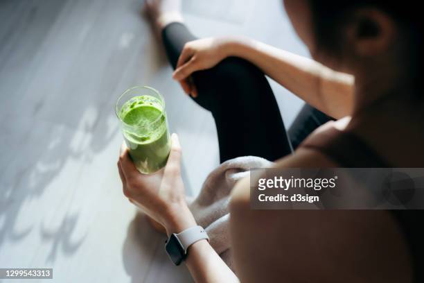 over the shoulder view of young asian sports woman taking a break, refreshing with healthy green juice after fitness work out / exercising / practicing yoga at home in the fresh bright morning - refreshment foto e immagini stock