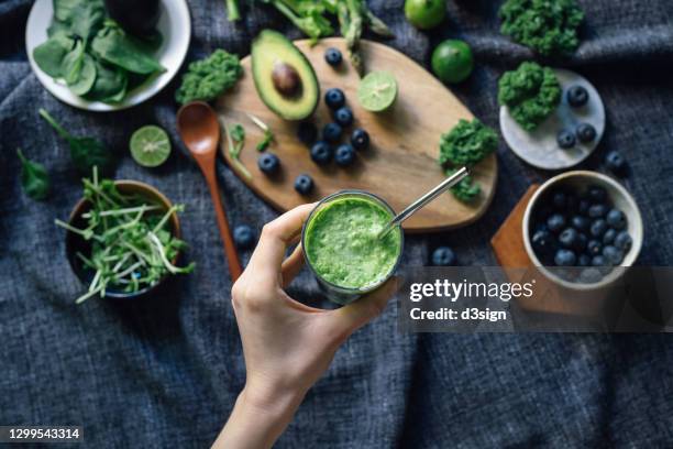 flat lay of fresh and healthy green fruits and vegetables on a rustic wooden board, against dark grey fabric for a healthy eating diet. woman's hand holding a glass of green smoothie with reusable straw. healthy superfood, green colour, detox diet concept - スムージー ストックフォトと画像